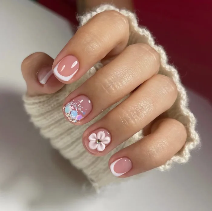 Short rounded nails with a sheer pink base featuring mixed accents: white French tips, chunky glitter, and a 3D white flower.