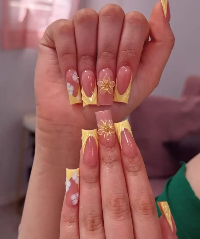 Long coffin-shaped nails with bright yellow French tips showcasing a unique ruffled smile line, accented with golden and white 3D flower embellishments.