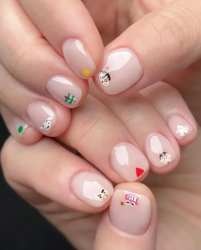 Very short rounded nails with a sheer nude polish base, each featuring a different tiny, colorful cartoon character or icon decal near the tip.