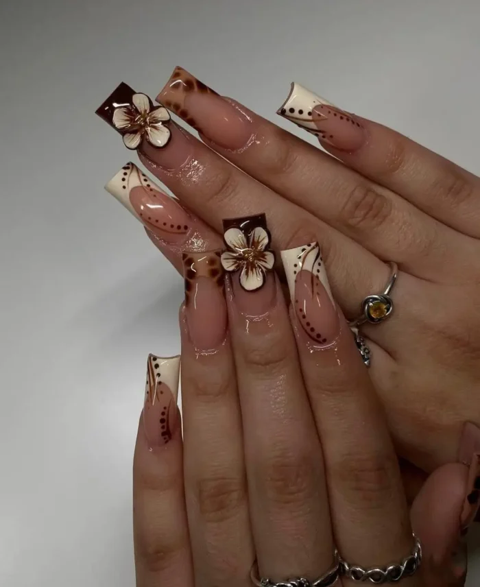 Long square nails featuring a modern French manicure with chocolate brown and cream angular tips, detailed with tiny brown dots and adorned with 3D cream and brown flower charms.