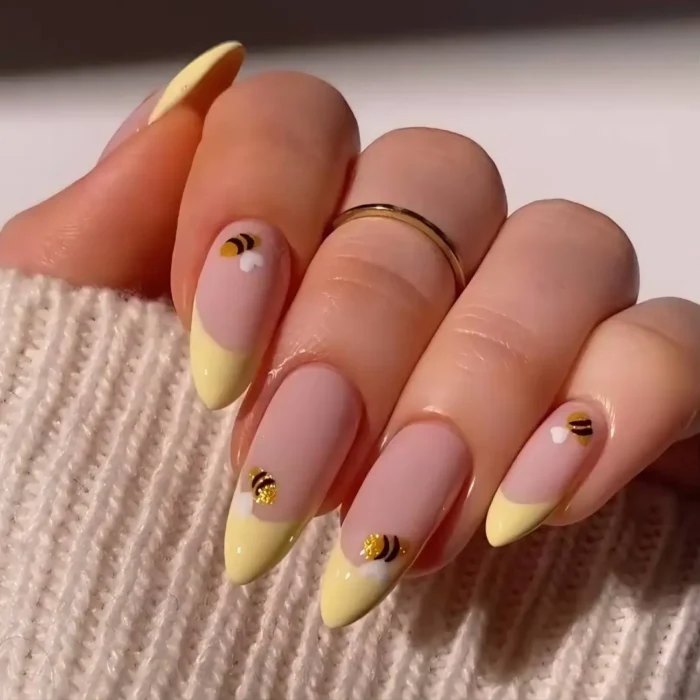 A close-up of long, almond-shaped nails featuring a butter yellow French manicure with small honey bee accents.