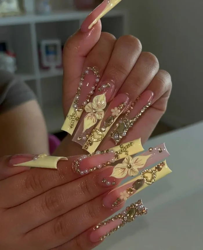 Extra-long square nails with pink to yellow ombré, adorned with 3D cream flowers, gold charms, pearls, and rhinestones.