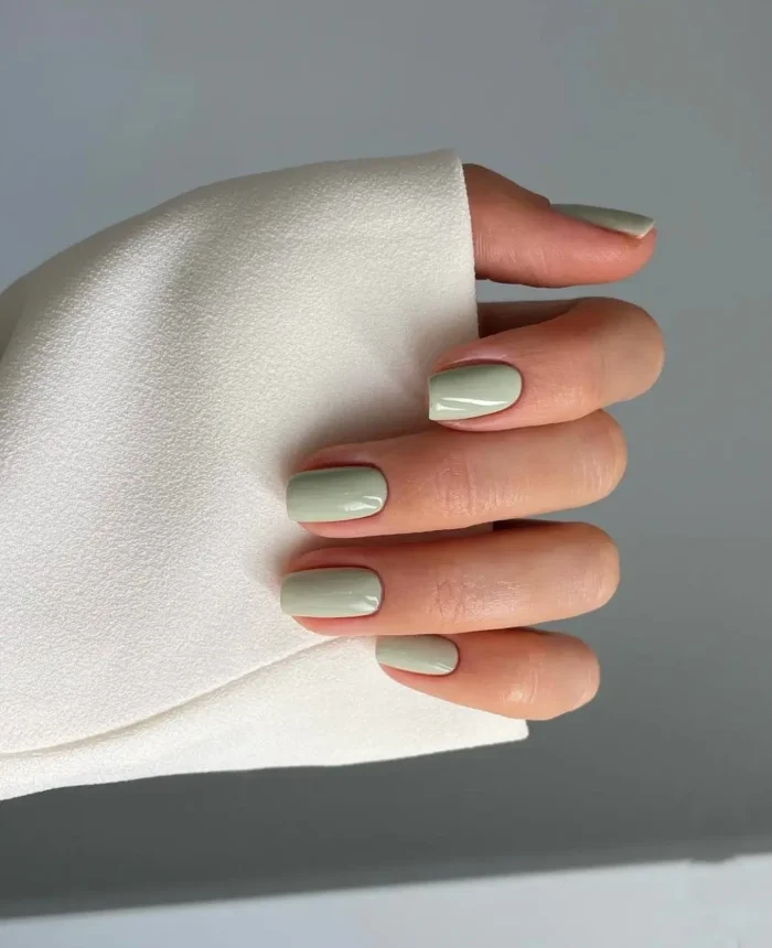 Muted sage green nail polish on neatly shaped short nails, showing a minimalist manicure.