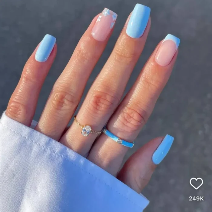 A hand with a mix-and-match manicure featuring sky blue nails, a blue French tip, and a sheer nail with tiny daisy nail art inspo.