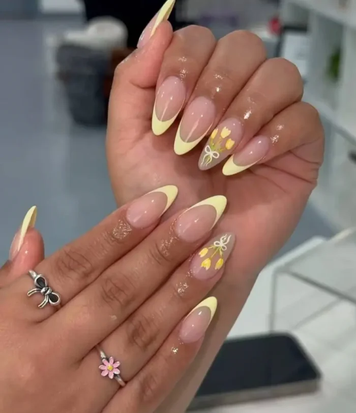 A butter yellow French manicure on almond nails, with an accent nail featuring hand-painted yellow tulips tied with a white bow.