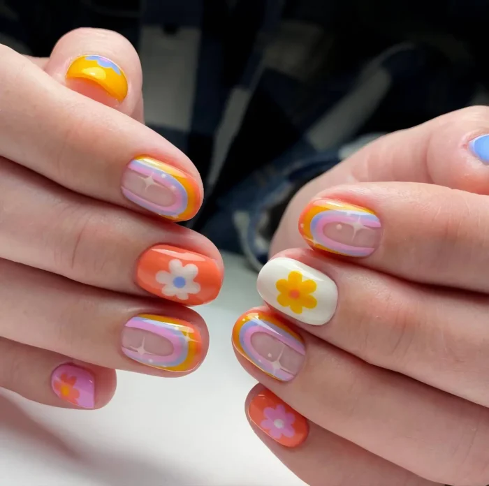 Short rounded nails with a colorful 70s retro theme featuring flower motifs, rainbow stripes, and wavy patterns in orange, yellow, and pink.