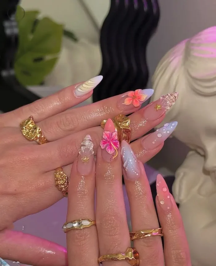 Long stiletto nails with a mermaidcore summer design, featuring 3D shells, flowers, pearls, and ethereal pastel accents.