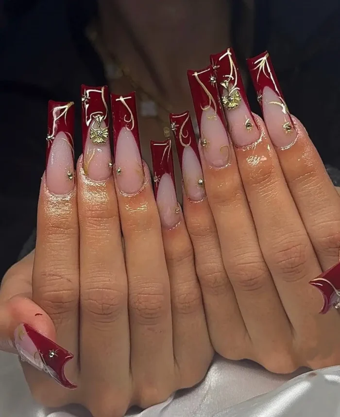 An ornate, long red French manicure with gold swirls, 3D bow charms, and rhinestone accents.