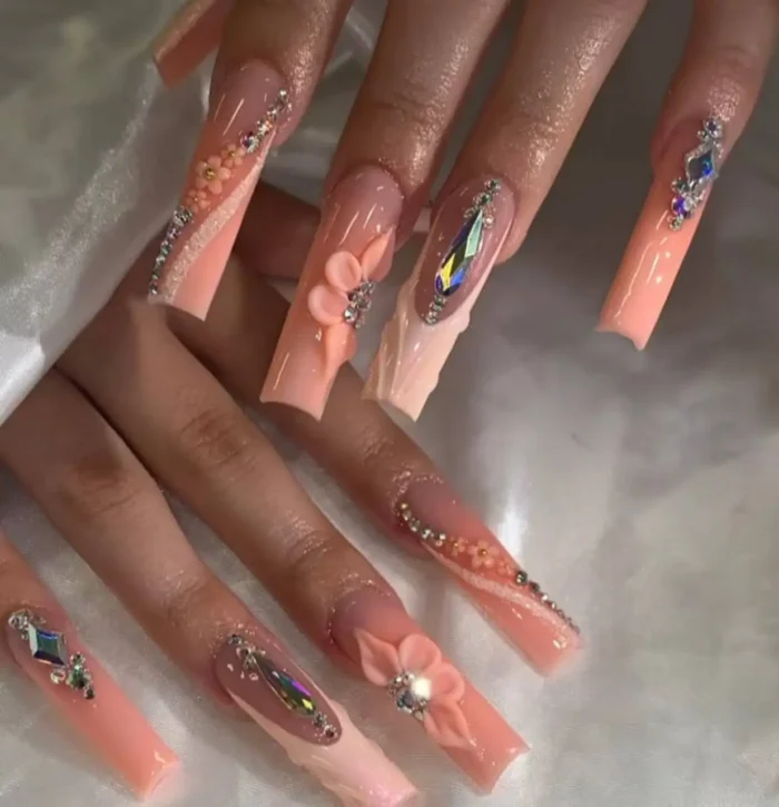 Long square nails in soft peach tones, adorned with 3D peach flowers and trails of iridescent crystals.