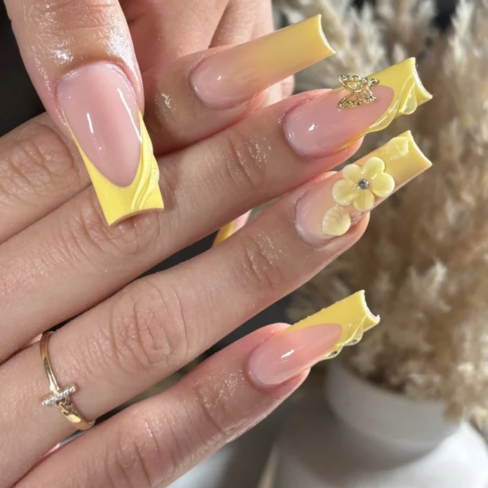 Long coffin-shaped nails with bright yellow sculpted French tips, accented with a 3D yellow flower and small gold charms on a sheer pink base.