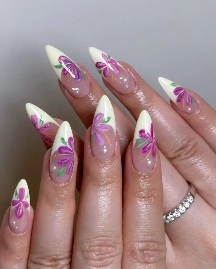 Long stiletto nails with white French tips featuring vibrant purple floral designs and pearl accents over a sheer base.