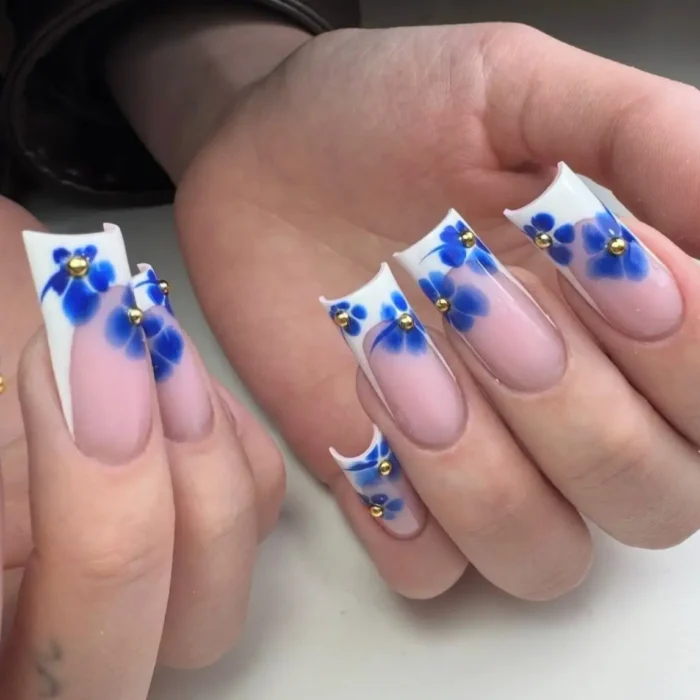 Long square nails with white angled French tips, adorned with royal blue watercolor-style flowers and gold bead centers.
