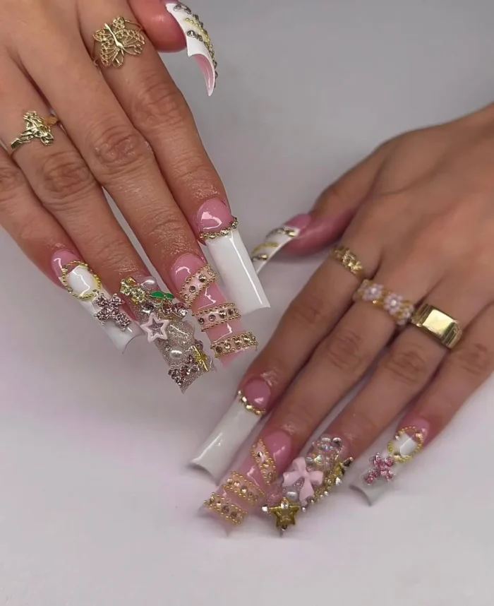 Extra-long white French tip nails lavishly decorated with gold bands, diverse 3D charms, and sparkling gemstones.
