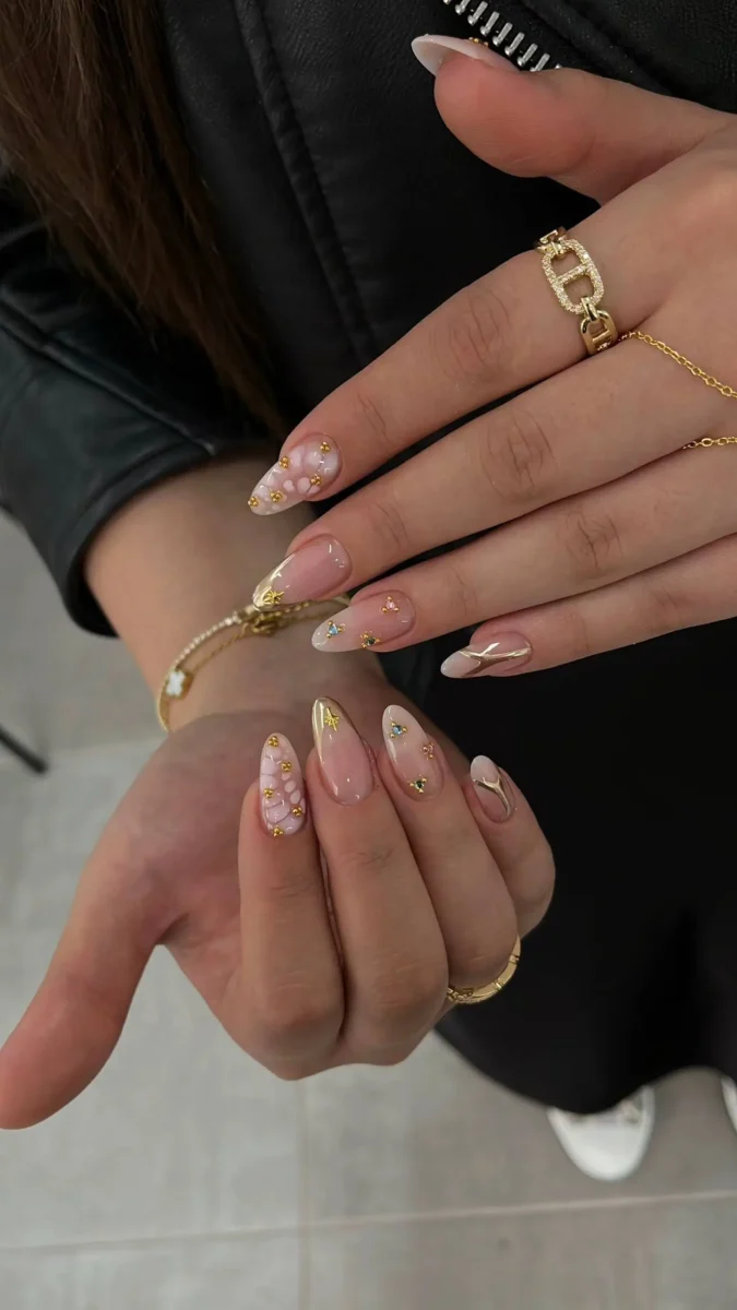 Almond-shaped nails with a milky blush pink base, featuring delicate scattered gold foil flakes, tiny colorful gemstones, and fine gold line accents.