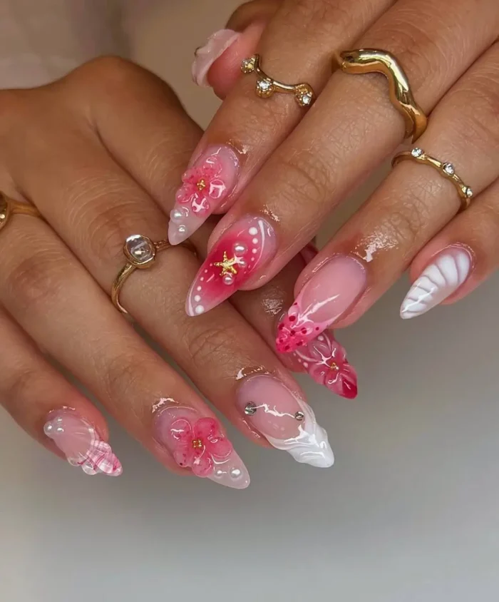 A maximalist pink and white manicure on long stiletto nails, featuring 3D flowers, pearls, gingham print, and a nail piercing.