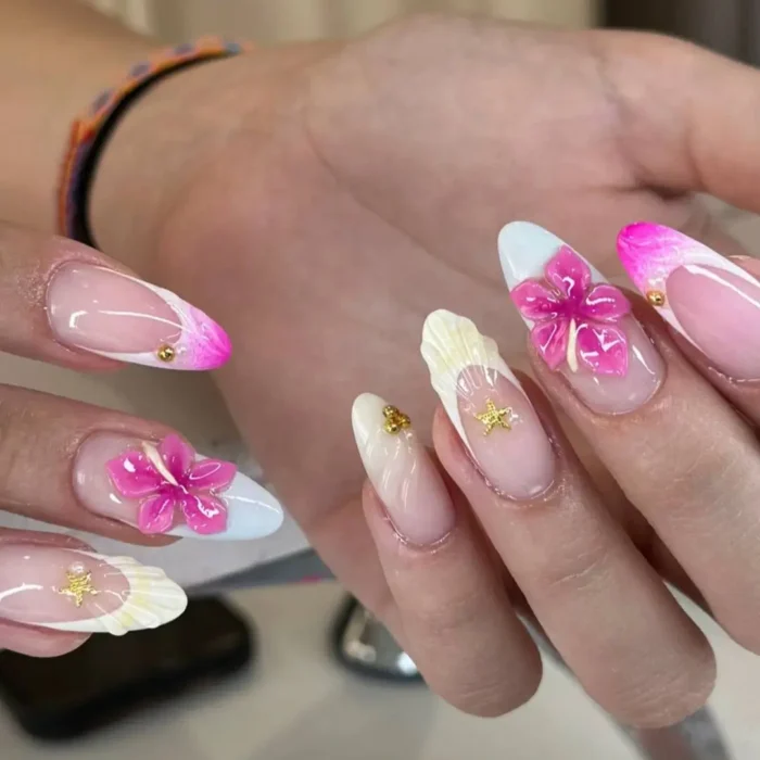 A tropical-themed manicure with vibrant pink 3D flowers, white accents, and seashell-textured nails with gold starfish.