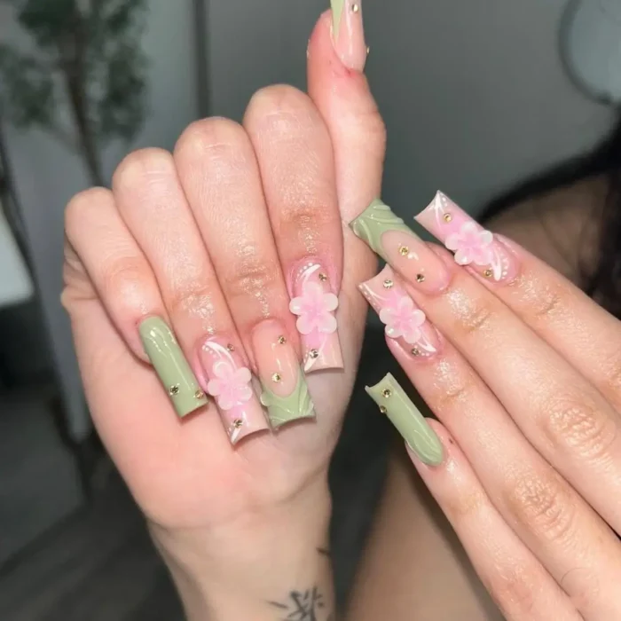 Long square nails with a mix of matte sage green polish and accent nails featuring 3D pink flowers and gold beads.