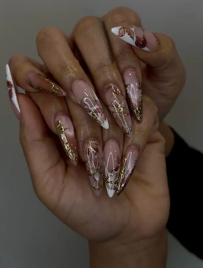 Long stiletto nails with a sheer base, featuring dimensional gold abstract rose designs, deep rose accents, and white details.