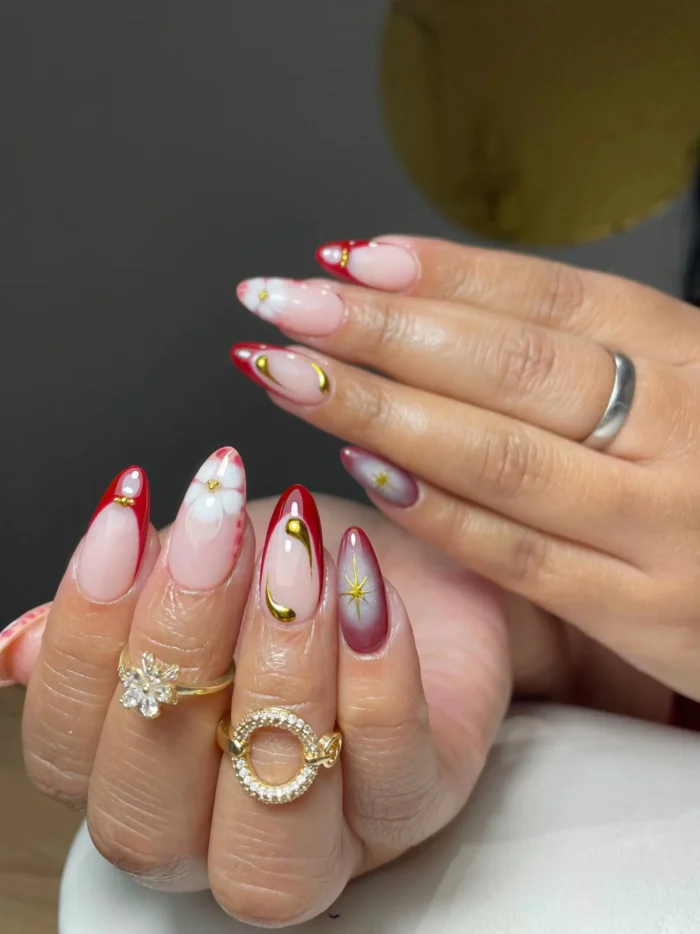 Long almond-shaped nails showcasing a mixed set of designs including red French tips, 3D white flowers, a red aura nail with a gold star, and abstract gold swirls.
