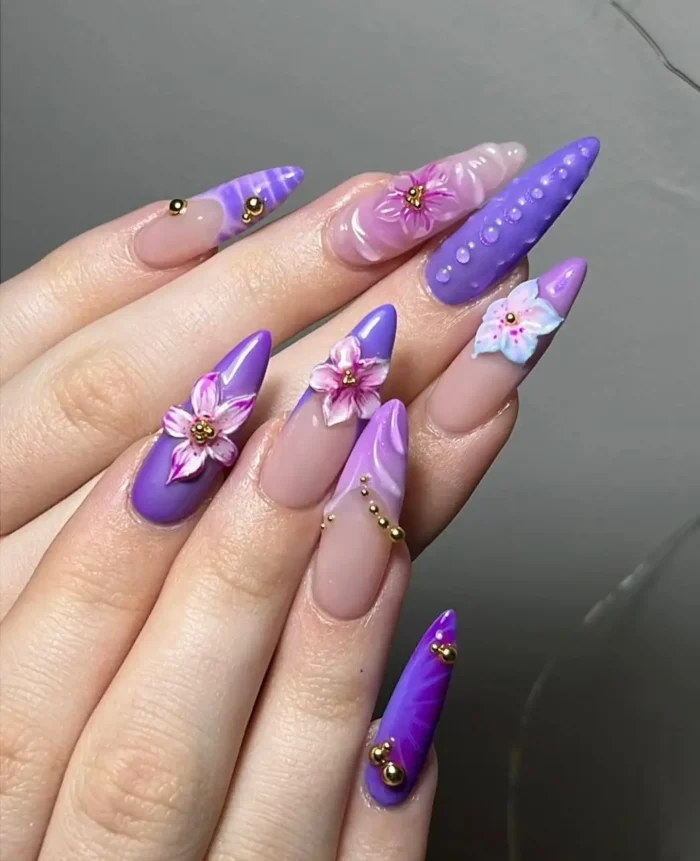 Pointed stiletto nails with a 3D violet vineyard purple nail design, adorned with purple and pink blossoms and gold beads.