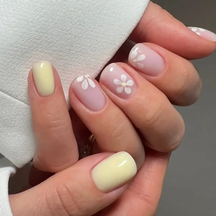 Short, rounded nails with a mix of pale yellow polish and sheer nude nails accented with white daisy designs.