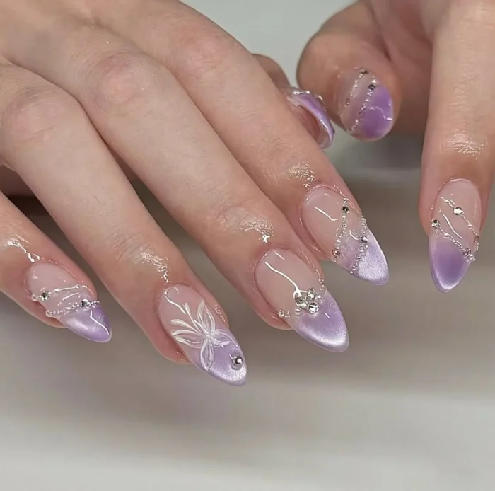 Long almond-shaped nails with shimmery lavender tips, detailed with delicate draped silver chain effects, tiny crystals, and a hand-painted white flower accent.
