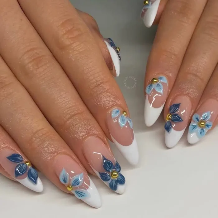 Almond-shaped French manicure with white tips, adorned with 3D blue flowers in various shades and small gold bead centers.