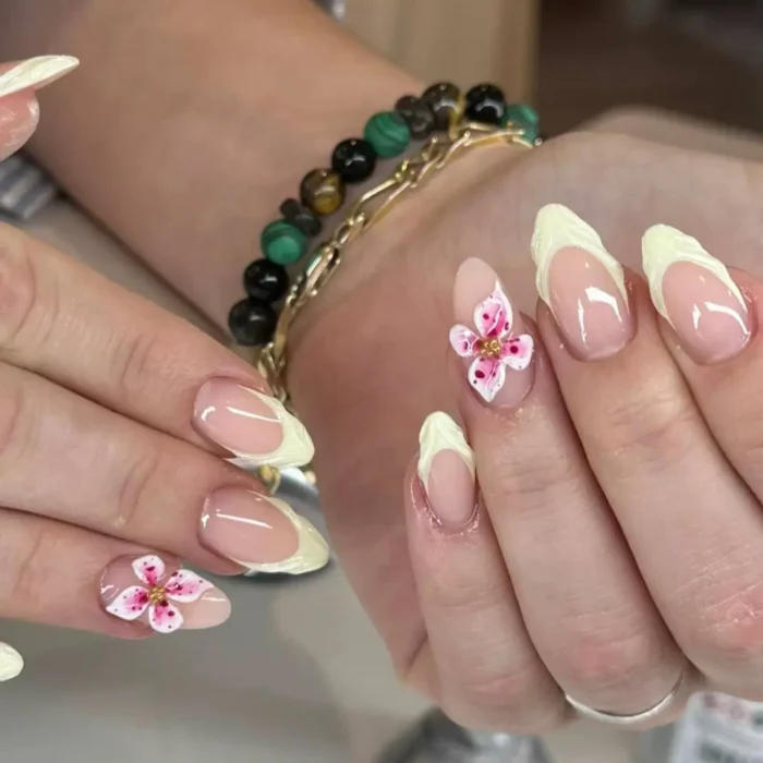 Almond nails with pale yellow French tips and accent nails featuring delicate 3D pink and white flowers with gold centers.
