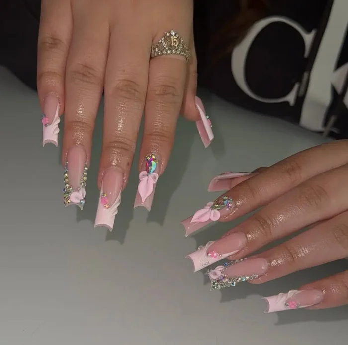 Long square nails with baby pink sculpted frill French tips, adorned with iridescent rhinestone clusters and large 3D pink bows.