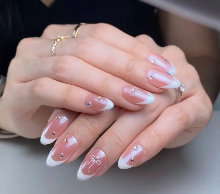 Almond nails with a dusty rose base and white French tips, accented with small gold studs and a white outline bow detail.