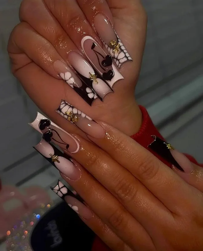 Long flared nails with black and white designs, featuring cherry motifs, spiderwebs, 3D white flowers, and gold stud accents.