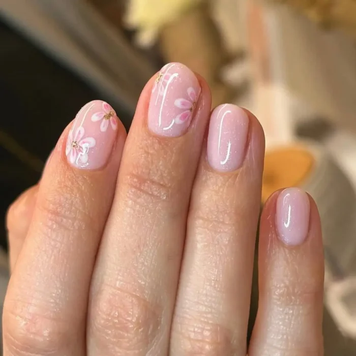 Short, rounded nails with a shimmery light pink base, adorned with delicate pink and white cherry blossom-style flowers with gold accents.