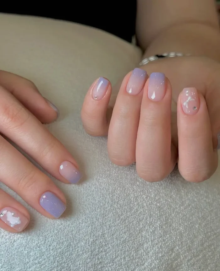 Short rounded nails with a mix of soft lavender-to-nude gradient, solid lavender polish, and sheer nude with white cloud designs and rhinestones.
