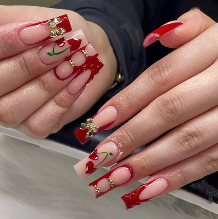 An elaborate red nail design with 3D jelly art, heart-shaped cherries, and large gold bow charms.
