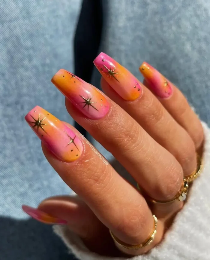 Long square nails with a pink-to-orange sunset ombre base featuring delicate gold starburst designs and gold stud accents.