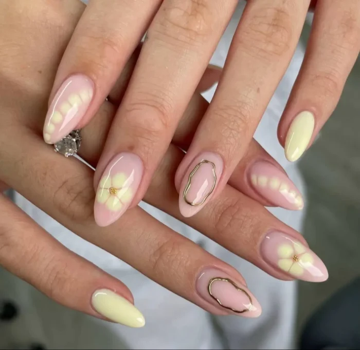 An elegant manicure on almond-shaped nails mixing solid butter yellow with 3D floral art, gold oval outlines, and petal-shaped butter yellow tips.