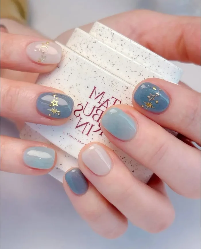 Cute simple gel manicure with muted blue and grey tones, featuring delicate gold star and moon celestial accents on short nails.