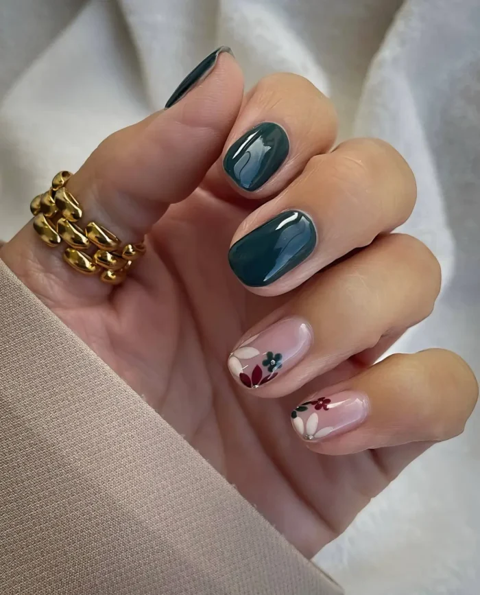Short rounded nails featuring solid deep emerald green polish mixed with sheer nude accent nails decorated with simple folk-art style flowers.