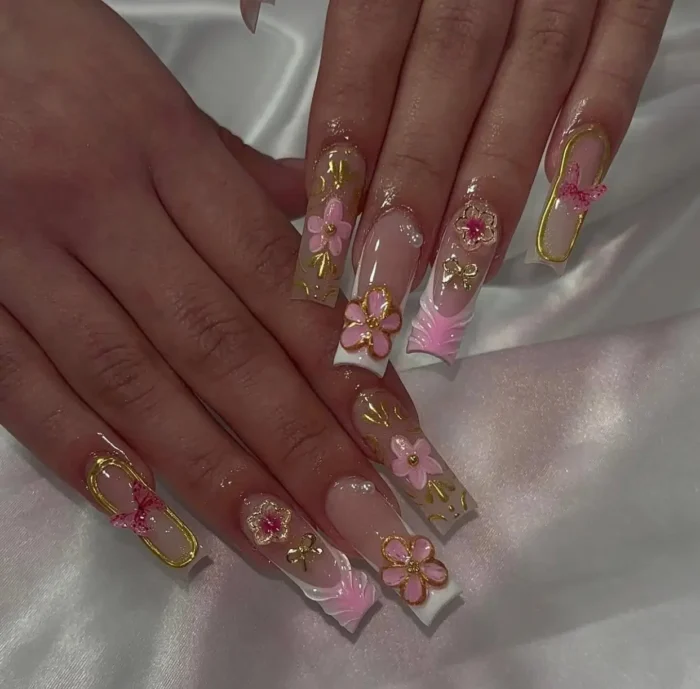 Very long square/coffin nails with a pink and gold theme, featuring textured French tips, 3D pink flowers, bold gold outline frames with pink bow charms, and other gold accents.