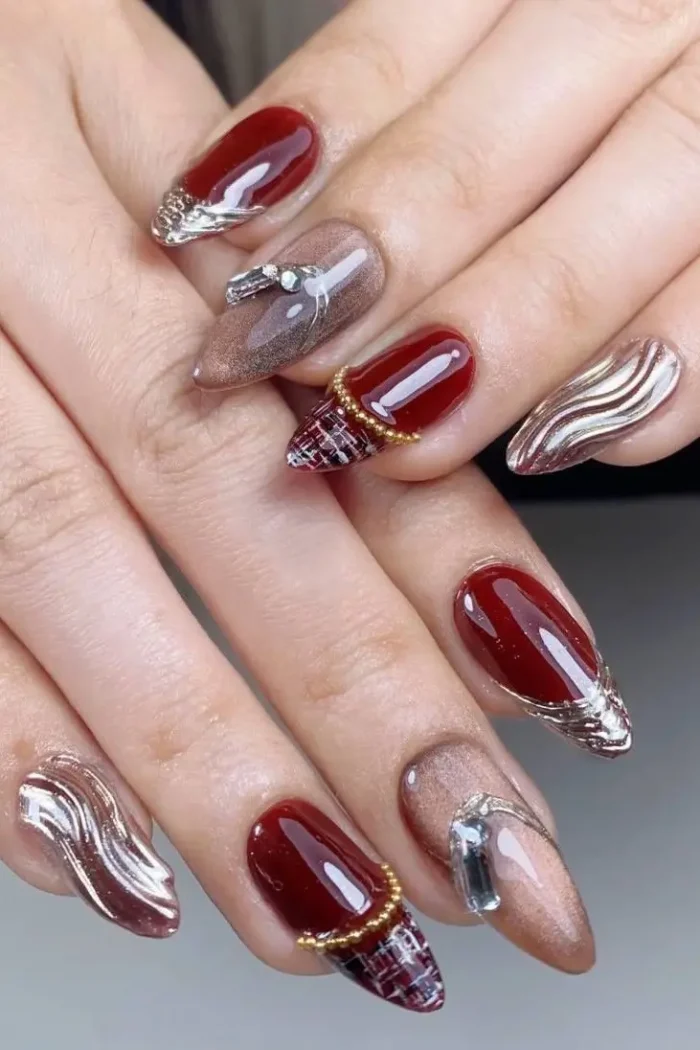 Long almond nails with a luxurious mix including glossy red, shimmery taupe, textured silver waves, red plaid tips with gold beads, and rhinestone accents.