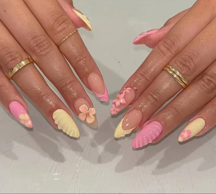 A vibrant manicure on stiletto nails featuring a mix of butter yellow and pink, with 3D seashell textures, floral art, and French tips.