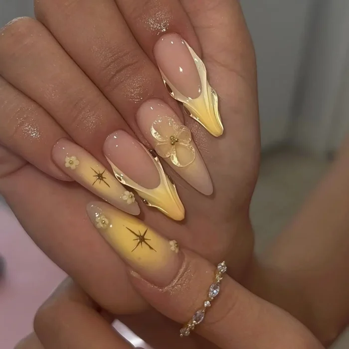 Long stiletto nails with yellow ombré and sculpted accents, featuring translucent 3D flowers and gold starburst details.