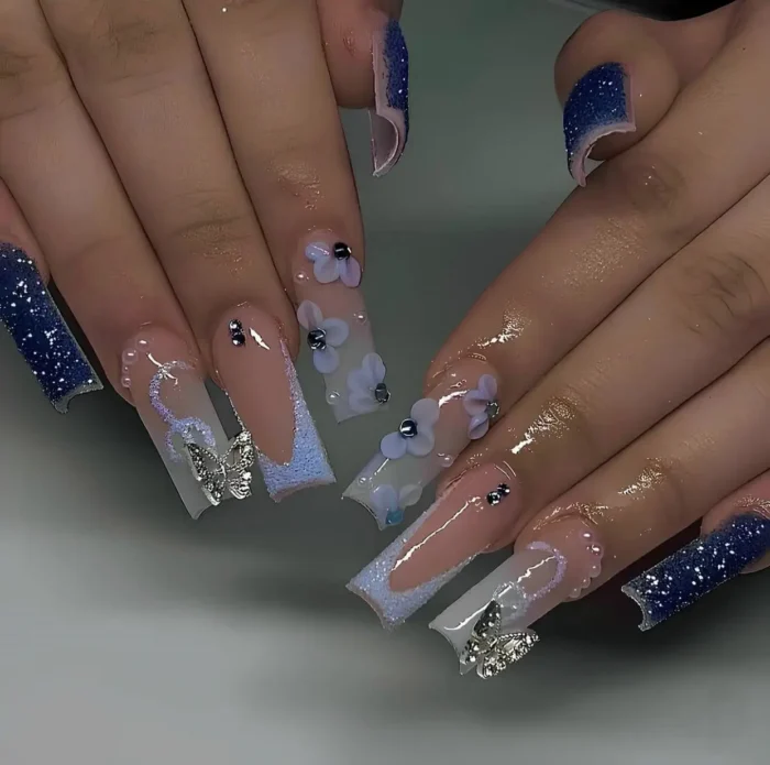 Long square nails with mixed midnight blue glitter and light blue sugared tips, silver butterfly charms, and 3D blue flowers.