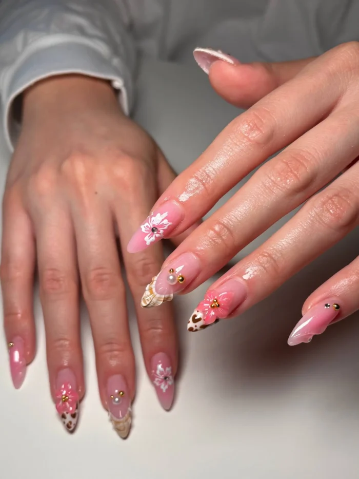 Almond-shaped nails with a mix of designs including subtle leopard print tips, soft pink blush, white 3D flowers, and delicate gold stud and pearl accents.