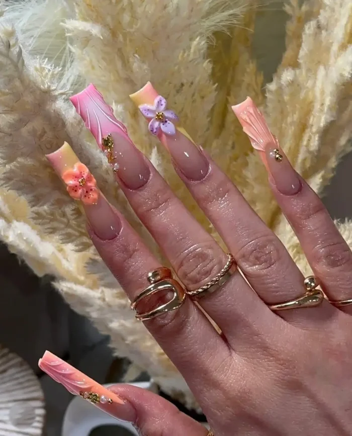 Artistic long square nails featuring a pastel ombre design in peach, yellow, and lavender hues, adorned with delicate 3D flowers and gold stud accents.