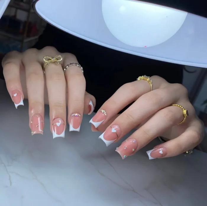 Medium-length square nails with a crisp white French manicure, featuring a charming heart-shaped cutout at the smile line and sparkling rhinestone accents.