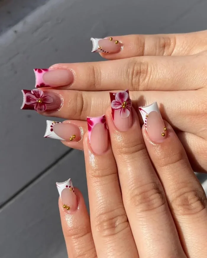 Square nails with deep French tips in white and pink, featuring large 3D pink floral accents with gold beads and gold stud details.
