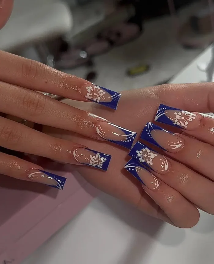 Long square nails featuring deep sapphire blue French tips, meticulously adorned with white hand-painted stylized flowers and ornate swirl detailing.