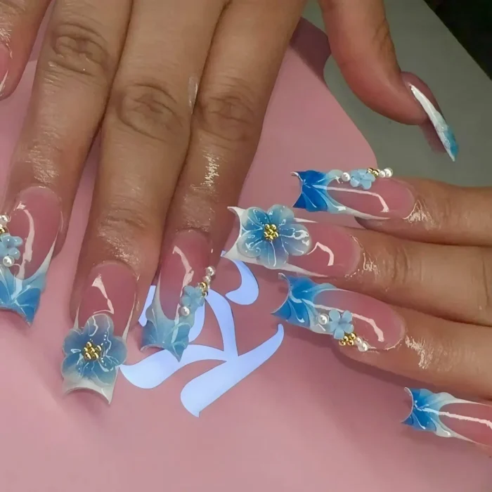 Long sculpted nails featuring intricate blue 3D flower designs, accented with pearls and gold beads.