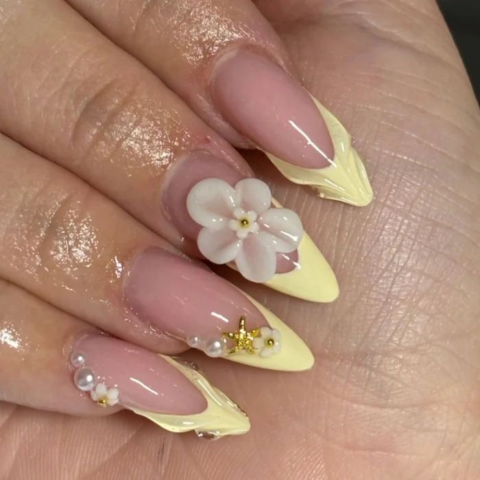Long stiletto-shaped nails with pastel yellow V-shaped French tips, embellished with 3D pink and white flowers, pearls, and tiny gold star accents.